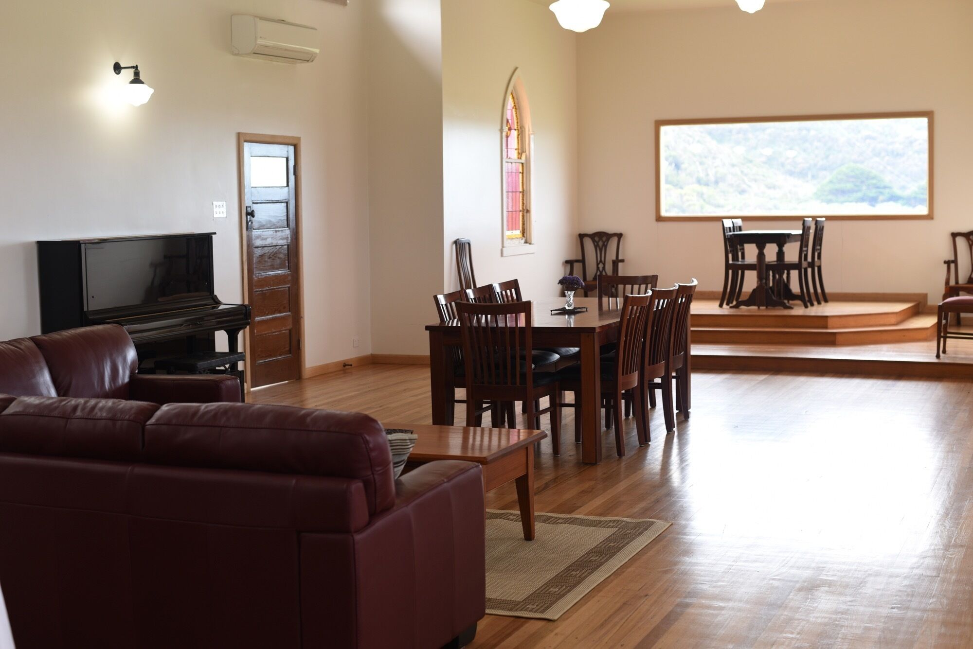 A spacious living area with a sofa, a dining table with chairs, a piano, and a large window with a view.