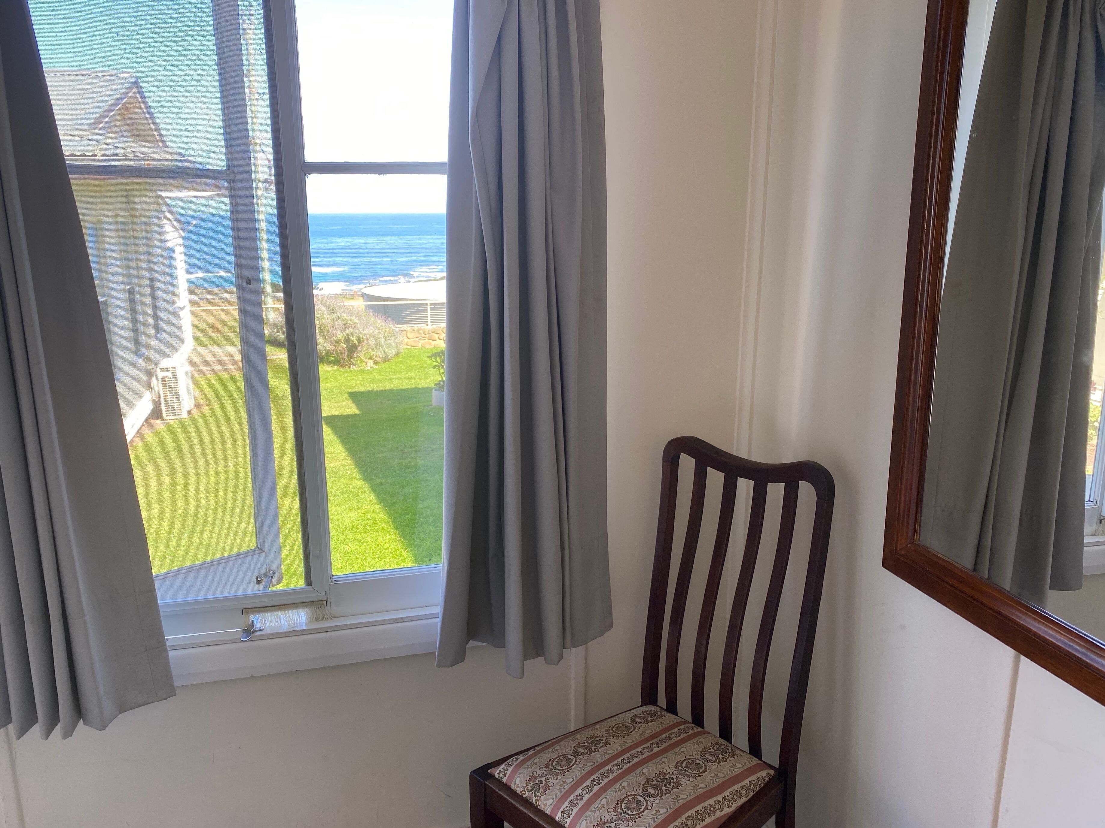 Una habitación con ventana que da a un área verde y un edificio. Hay una silla de madera con cojín estampado.