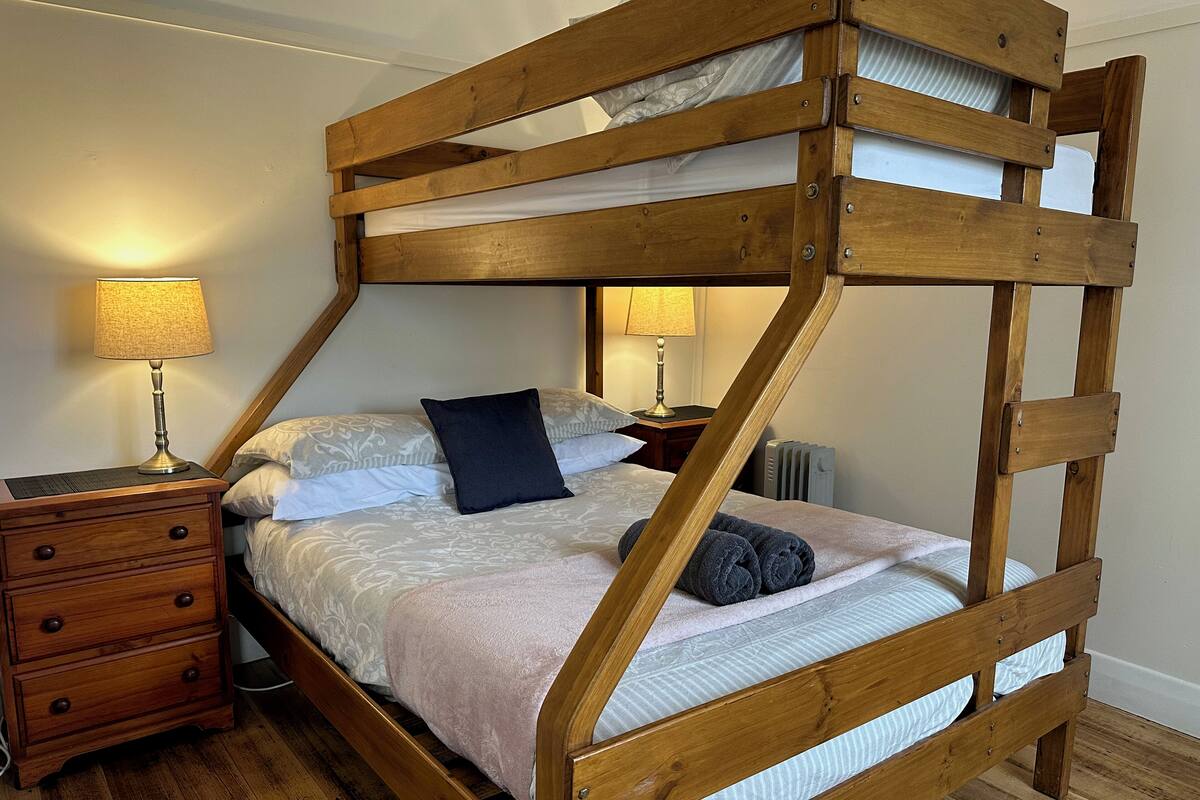 Una cama nido de madera con un mesita de noche y una lámpara, una almohada y una toalla enrollada sobre la cama.