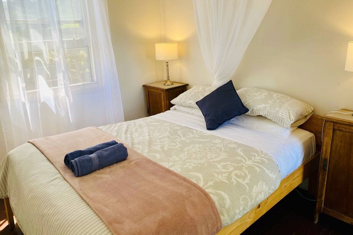 Una cama con una colcha beige y blanca de estampado, un almohán azul y una manta marrón. Hay dos mesitas de noche con lámparas, y una ventana con cortinas traslúceras.