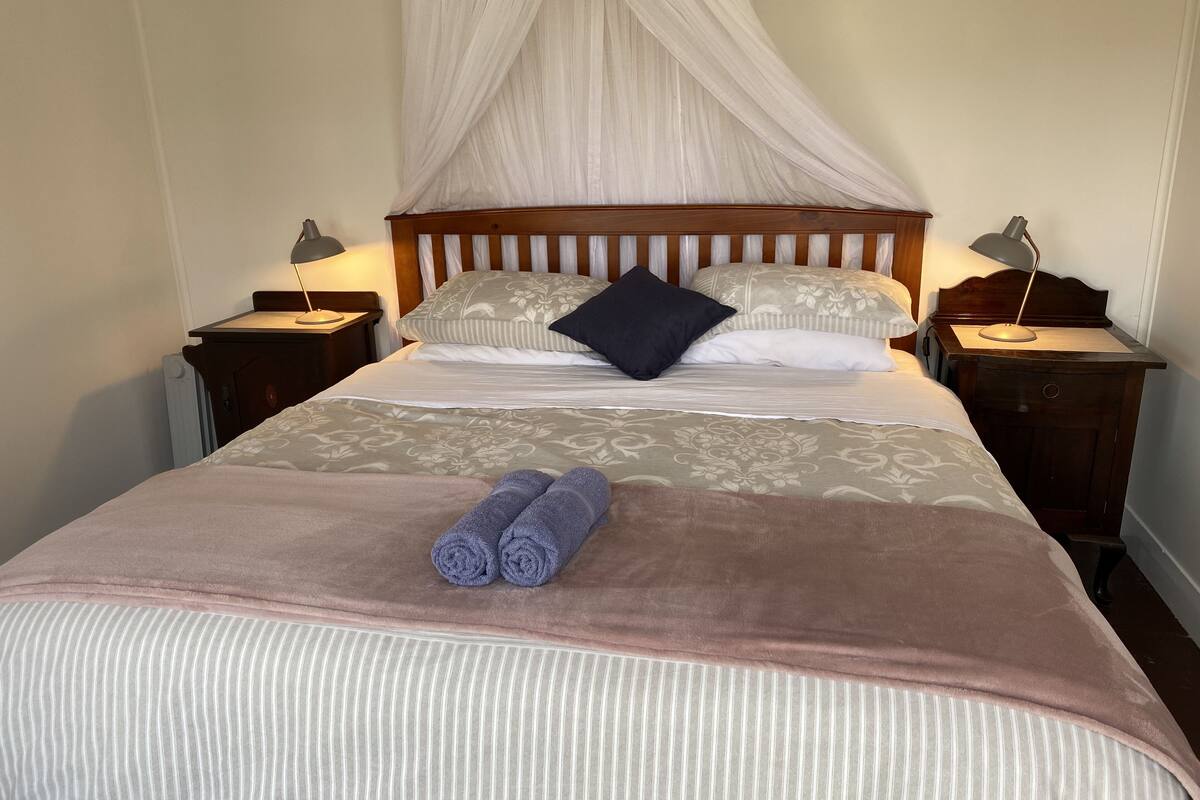 Una cama bien hecha con dosel, dos mesitas de noche con lámparas y toallas enrolladas sobre la cama.