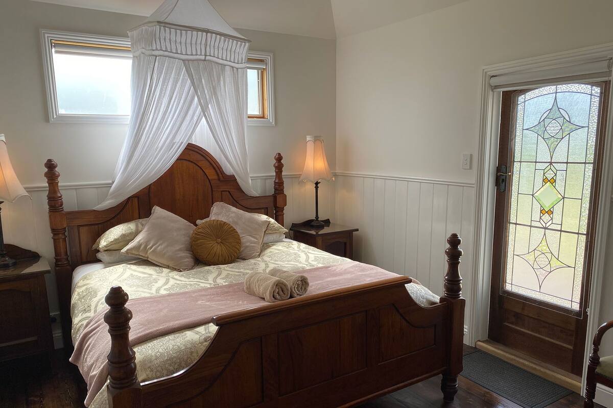 Un dormitorio con cama con dosel, dos mesitas de noche con lámparas, suelo de madera y puerta de vitral.
