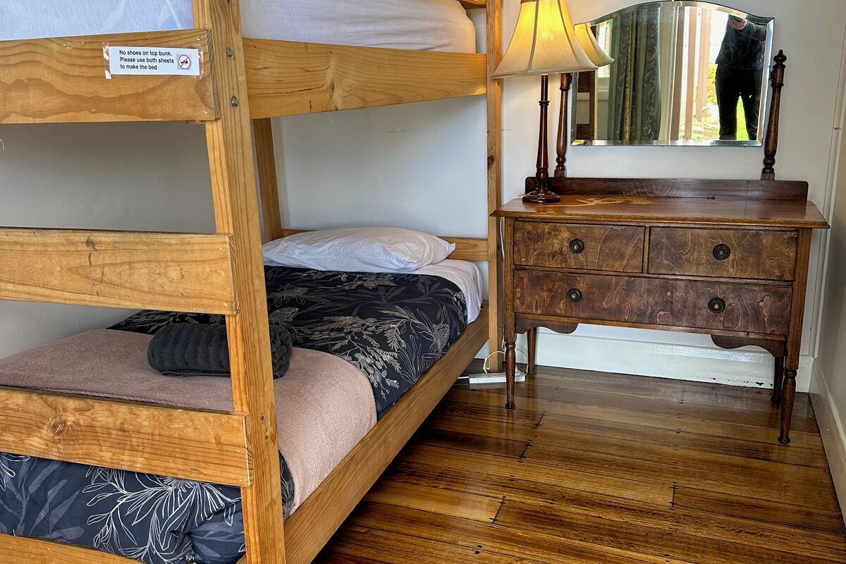 Una cama litera con un letrero que indica no poner zapatos encima. Una cómoda de madera con cajones y un espejo están junto a la cama.