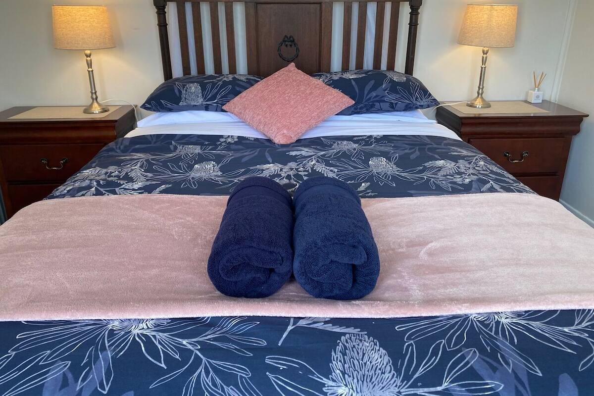 Una cama bien tendida con un edredón estampado con motivos florales, una manta rosa y dos toallas enrolladas.
