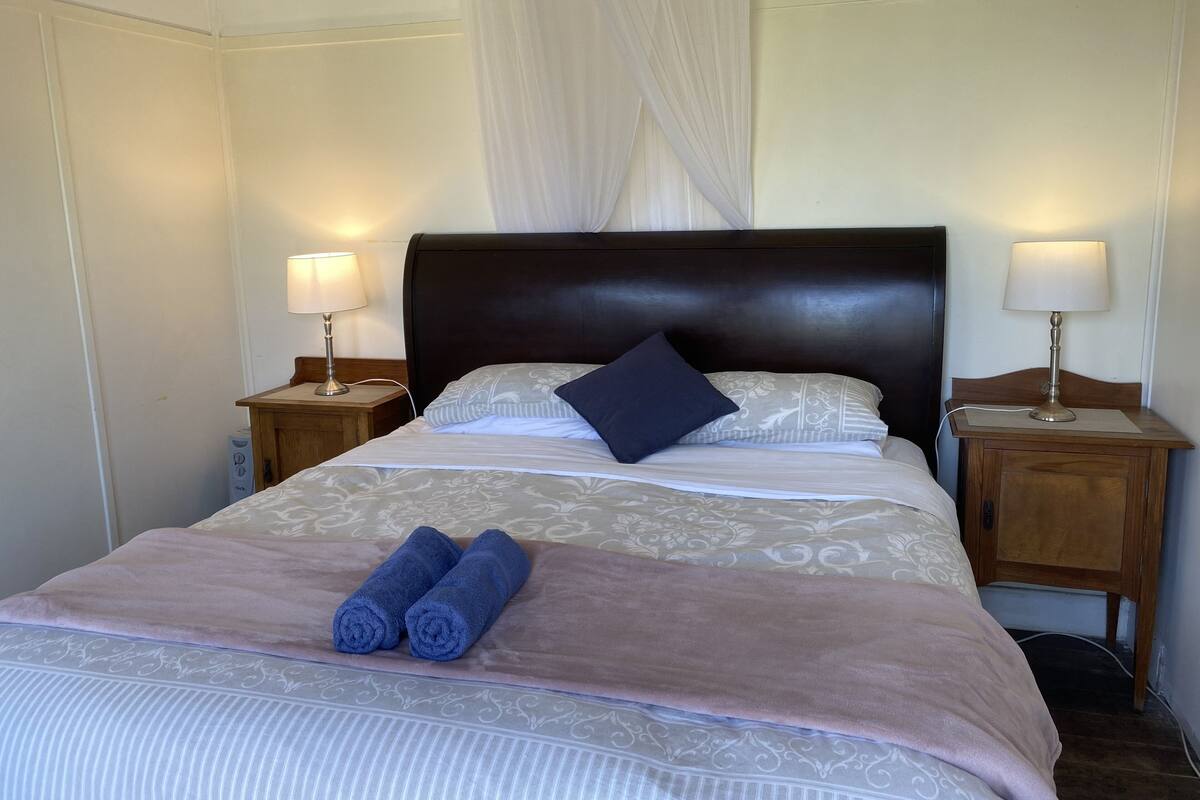 Una cama bien hecha con cabecera, dos lámparas de noche y toallas enrolladas sobre la cama.