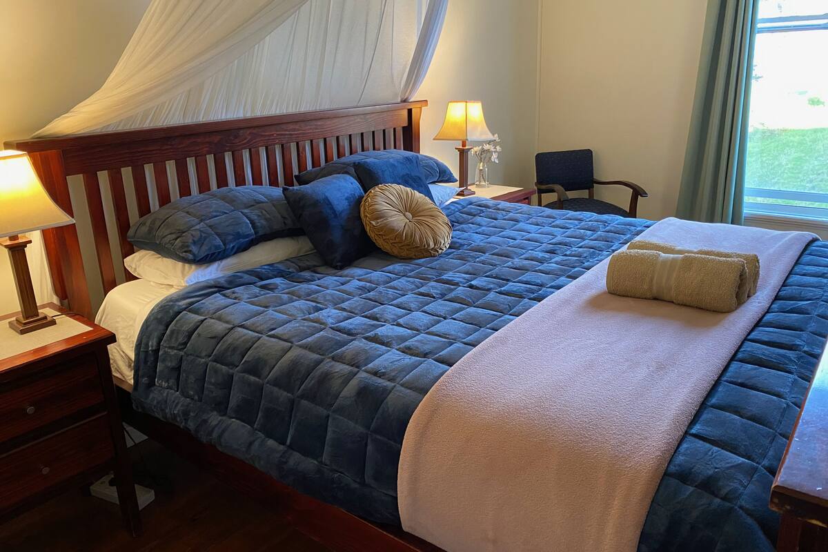 Una cama bien hecha con un edredón azul, una manta blanca y una toalla color beige. Hay una mesita de noche con una lámpara y una pequeña mesa con una lámpara y un jarrón de flores.