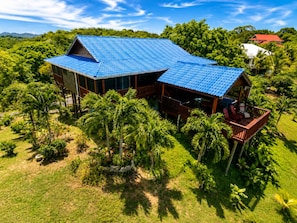 Una casa grande con techo azul, rodeada de abundante vegetación y palmeras.