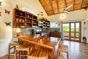 Une cuisine dotée d’un îlot central, de tabourets de bar, d’étagères en bois et d’une vue sur l’extérieur grâce à une porte vitrée.
