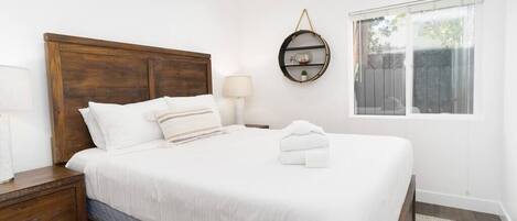 A neatly made bed with white linens, a wooden headboard, a bedside lamp, and a small round shelf with decorative items.