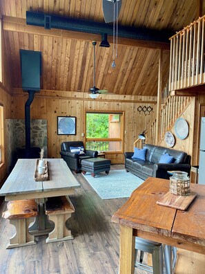 A wooden cabin interior with a dining table, two sofas, a fireplace, and a large window.