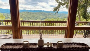 A wooden table with a woven tray, salt and pepper shakers, and a candle holder on a balcony overlooking a forested mountain range.