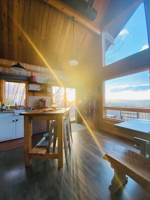 A cabin interior with a wooden dining table, a bench, a kitchen area with a sink and cabinets, and a large window offering a view of the outdoors.