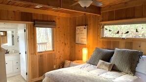 A wooden cabin bedroom with a bed, a ceiling fan, a window with a view of trees, and a sign on the wall.