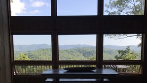 A room with a large window overlooking a forested mountain range. There is a wooden table and benches in the center.