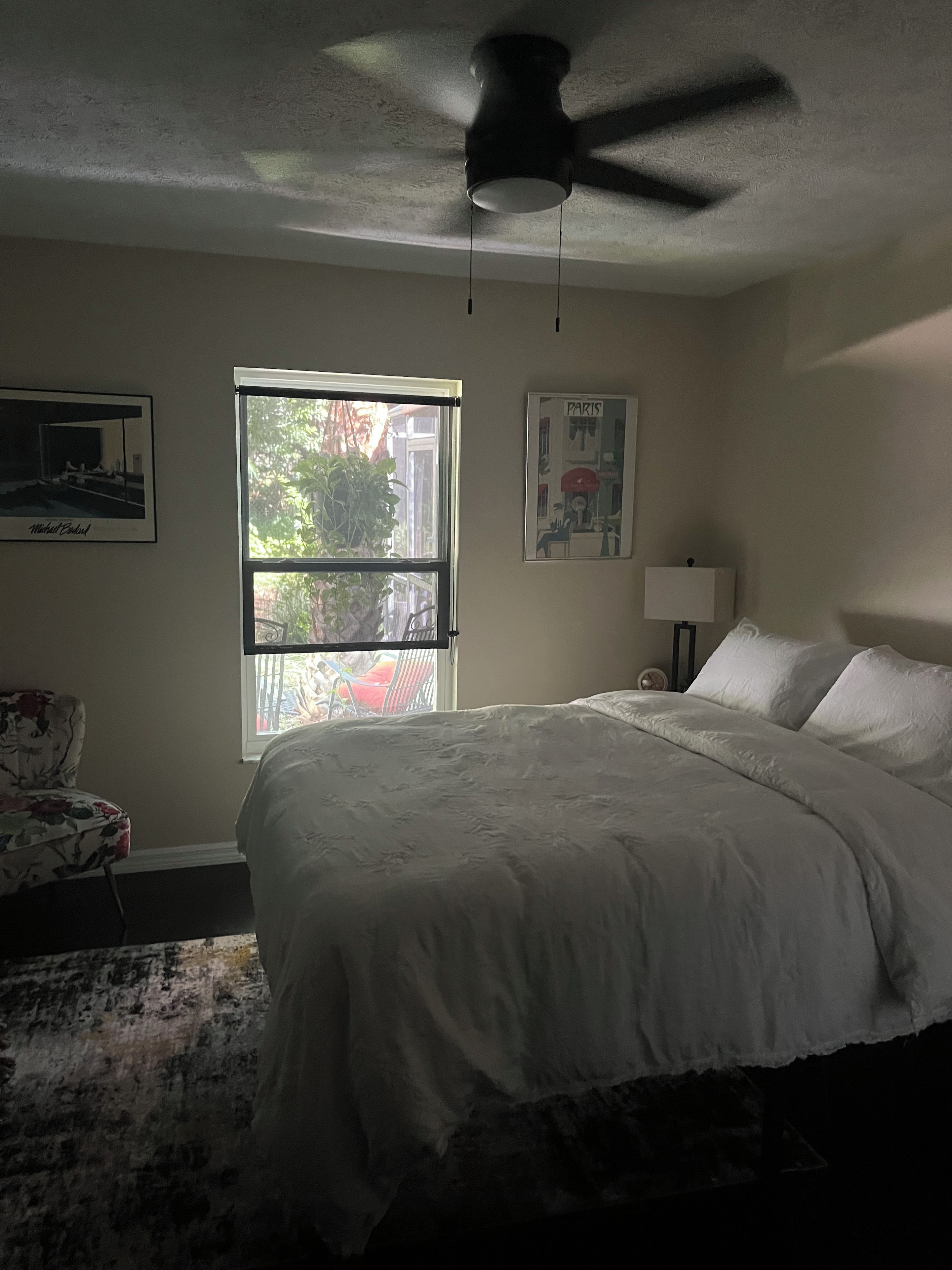 Une chambre à coucher avec un lit, un ventilateur de plafond, une fenêtre et un mur orné d’un tableau encadré.