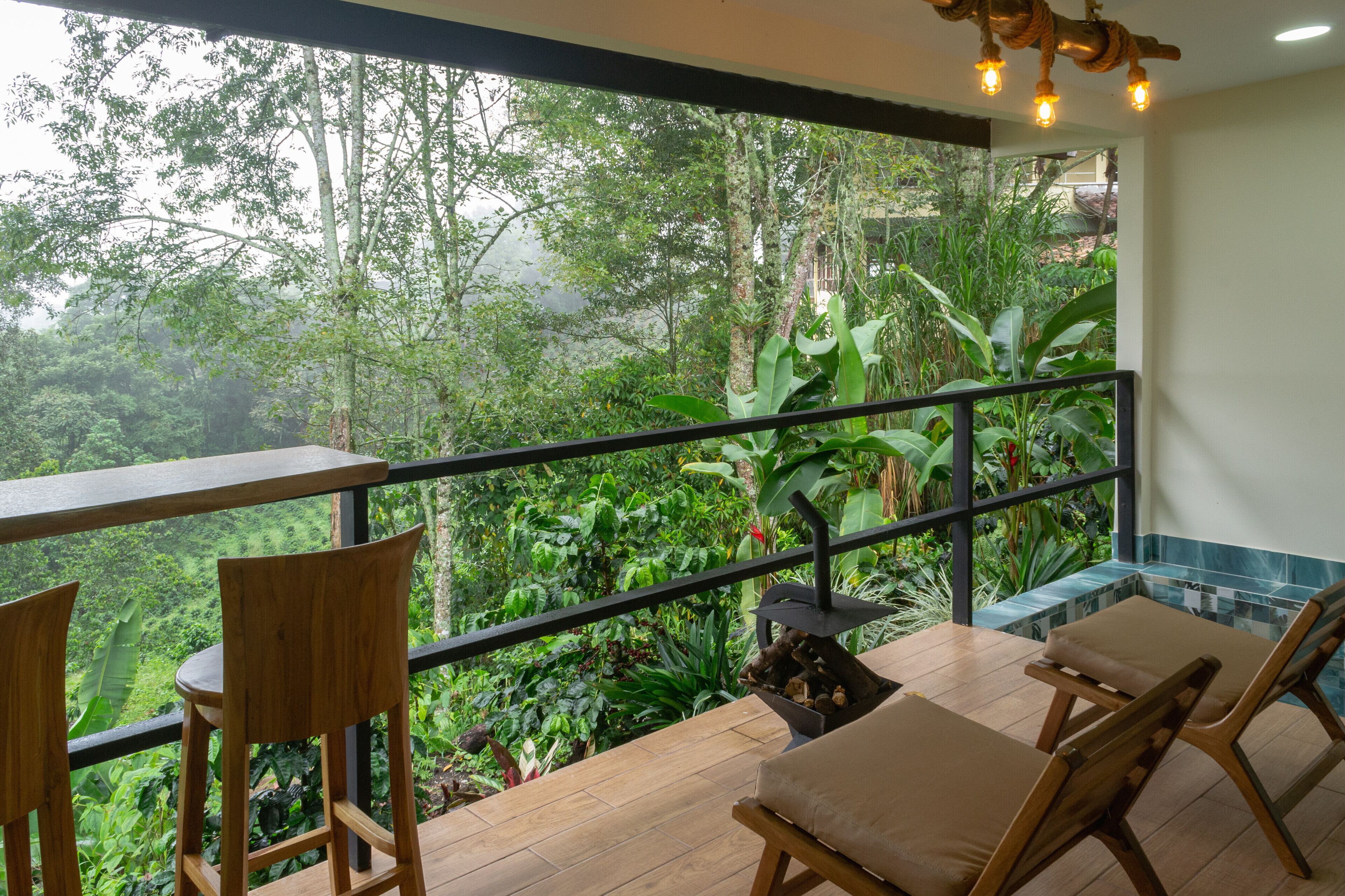 A balcony with wooden chairs and a table, overlooking a lush garden and forested area.