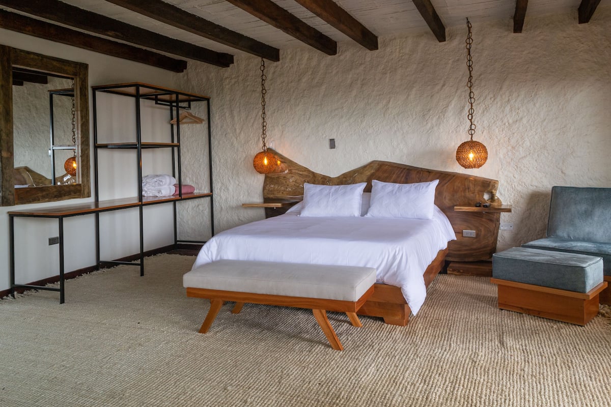 A bedroom with a wooden bed, a bench, a nightstand, a chair, a hanging light, and a mirror.