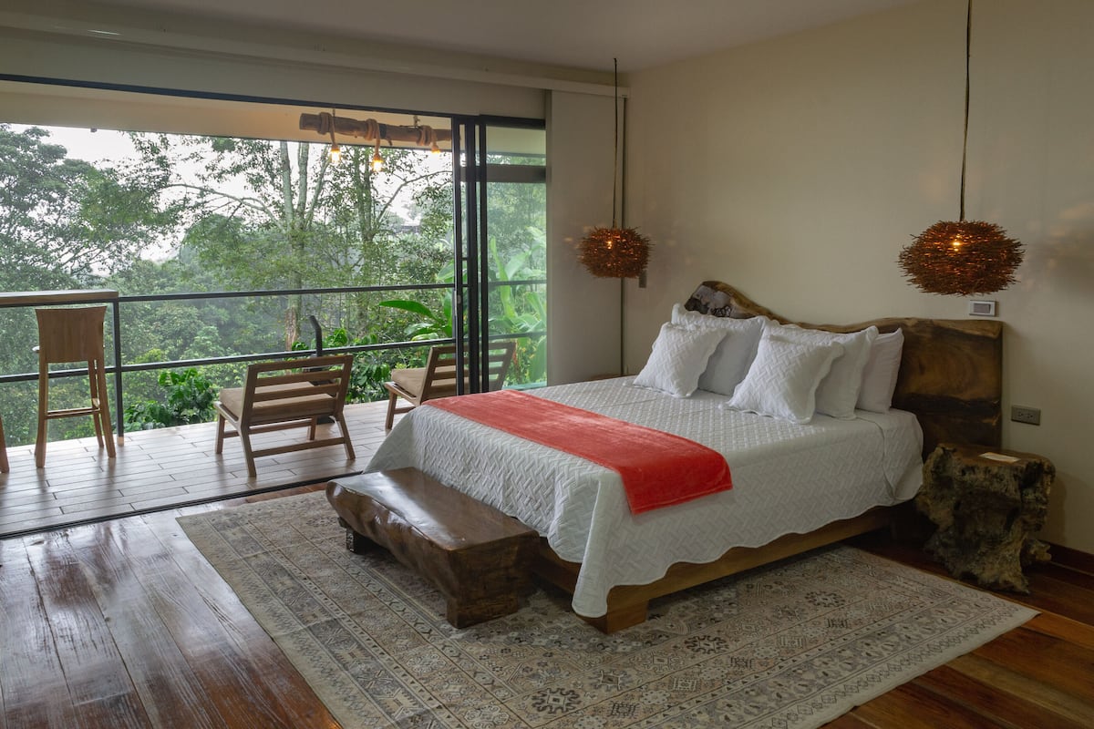 A bedroom with a large bed, wooden headboard, and a bench. There is a balcony with a view of greenery outside.