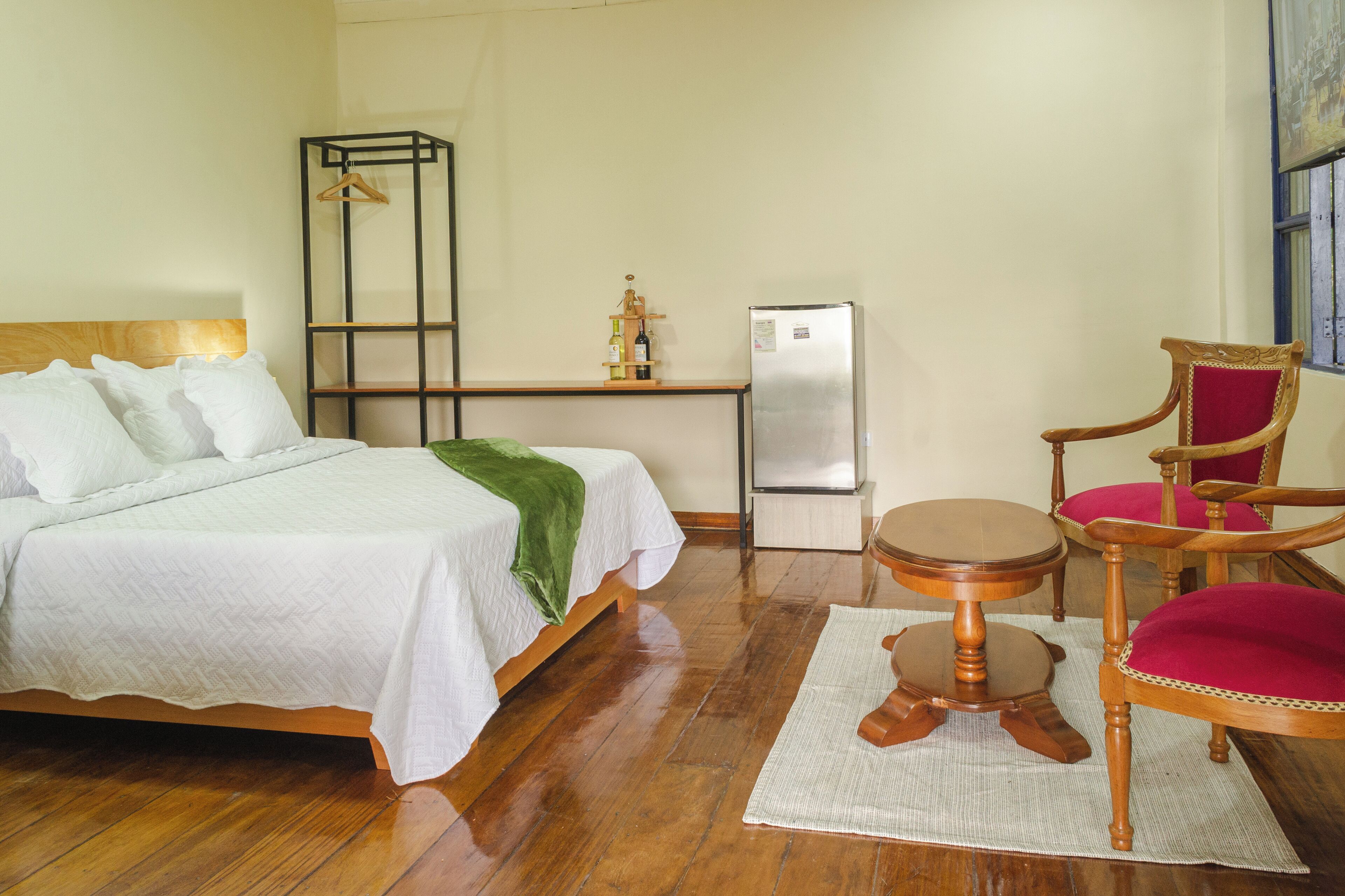 Habitación de hotel con una cama de madera, una mesita redonda pequeña, una silla, un armario y un refrigerador.