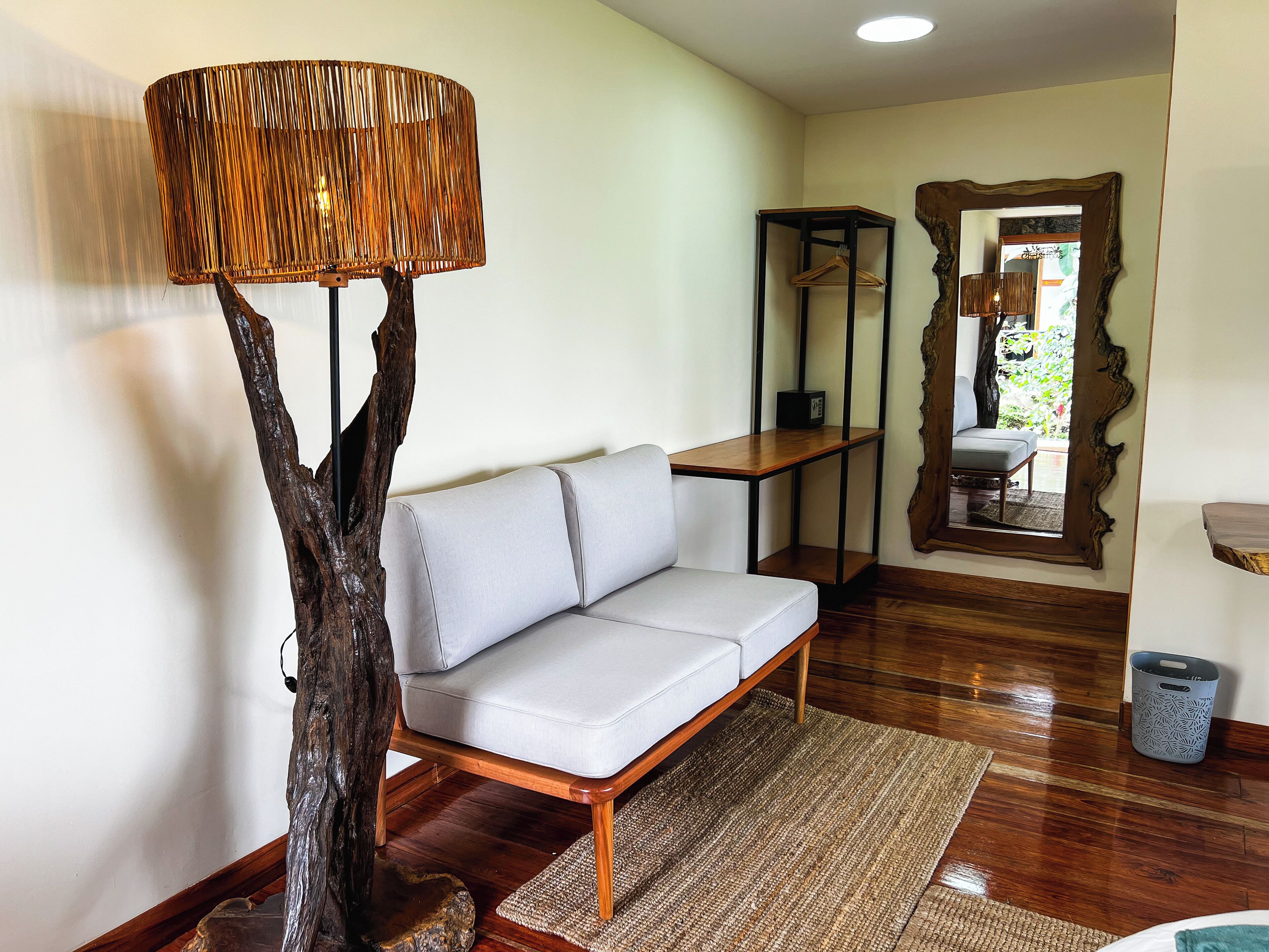 A modern living room with a wooden floor, a grey sofa, a unique tree-shaped floor lamp, a small table, and a mirror with an ornate frame.