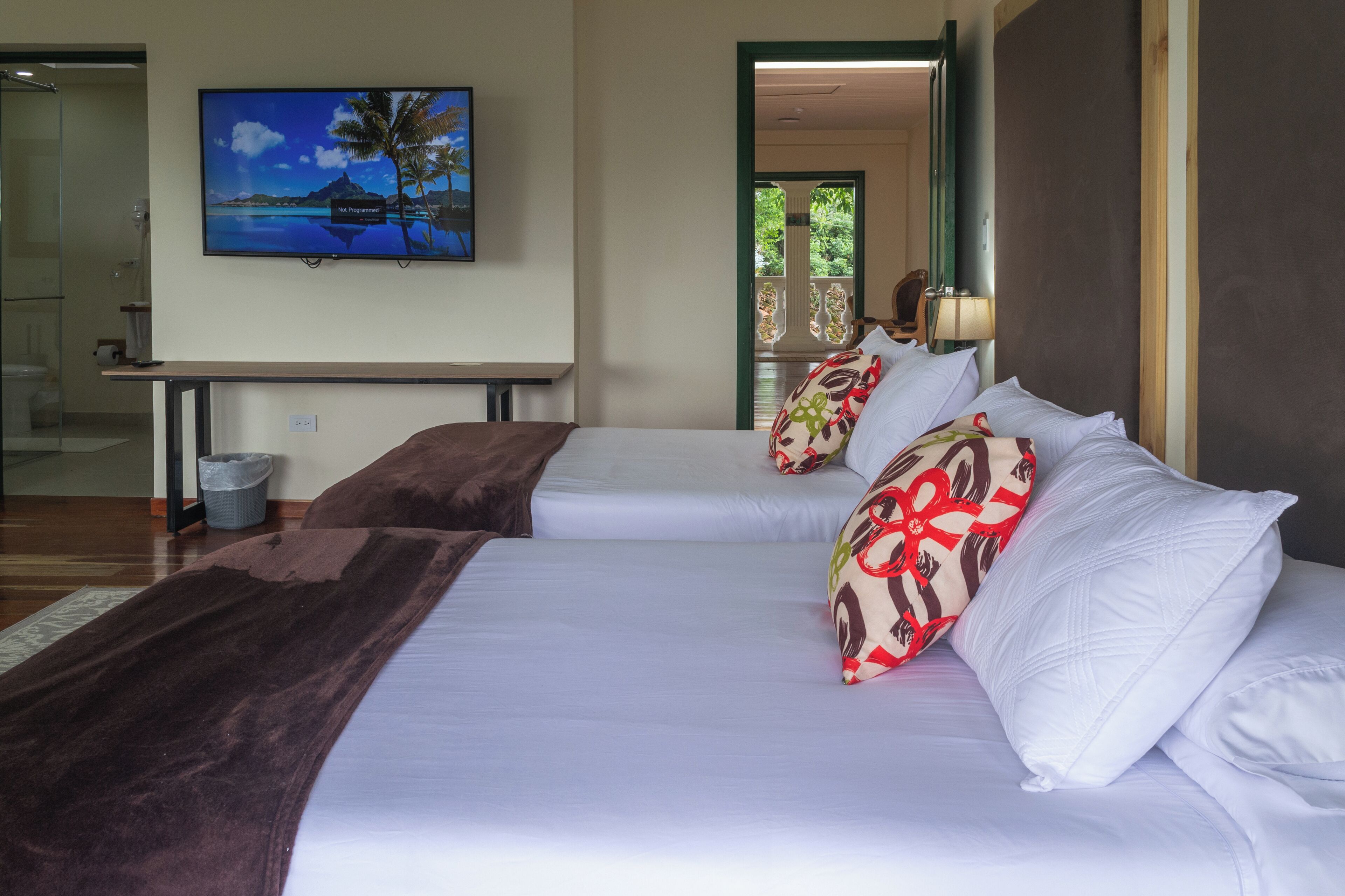 A hotel room with two beds, a television, and a view of a palm tree and water.