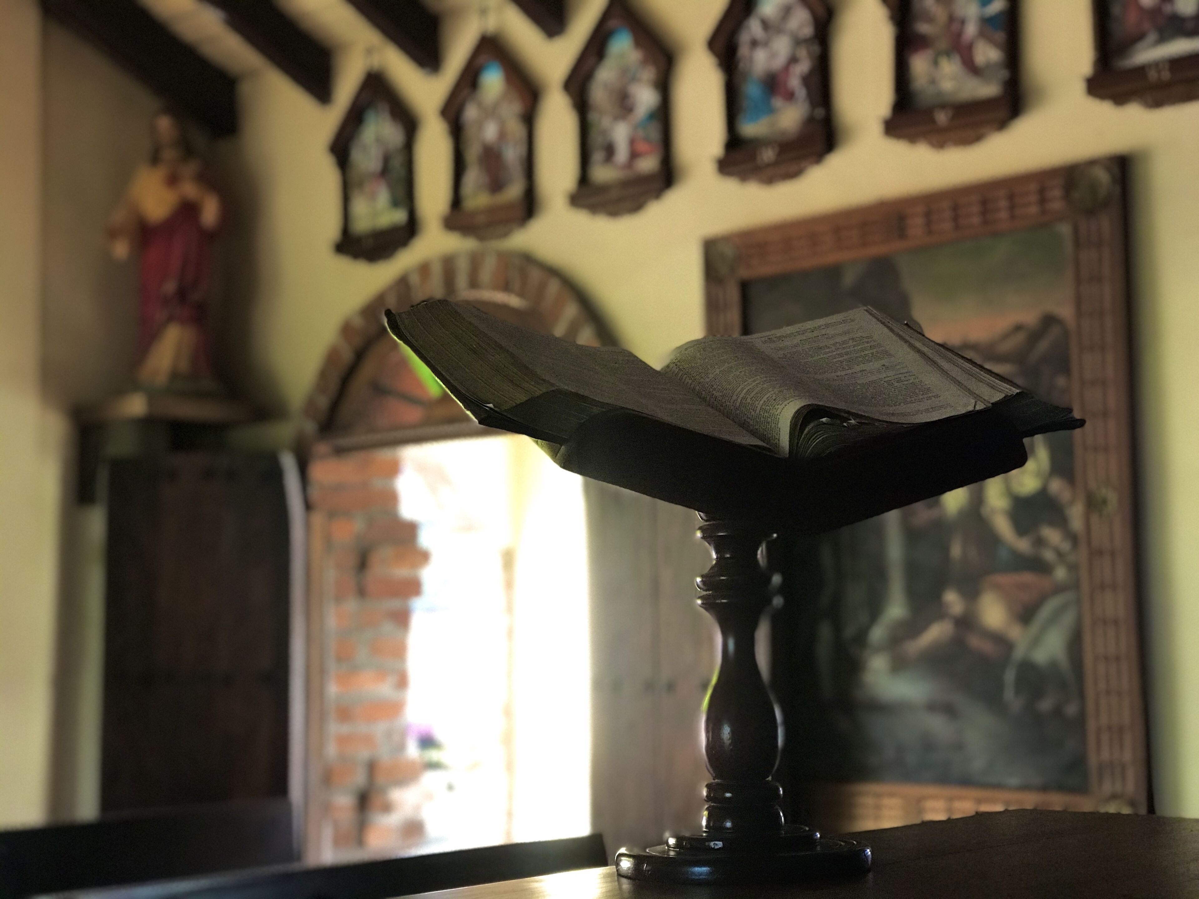 Un atril de madera con un libro abierto, una estatua y vidrieras en una habitación con paredes de ladrillo y un cuadro.