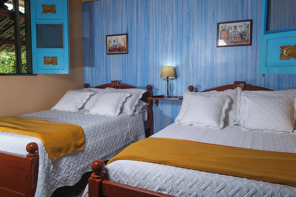 A room with two beds, a nightstand with a lamp, and a window with a view of greenery.