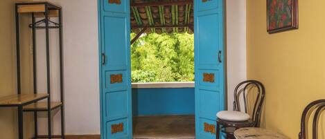 Une pièce dont la porte est ouverte et qui donne sur un balcon offrant une vue sur la verdure. On y trouve deux chaises en bois et une petite table.