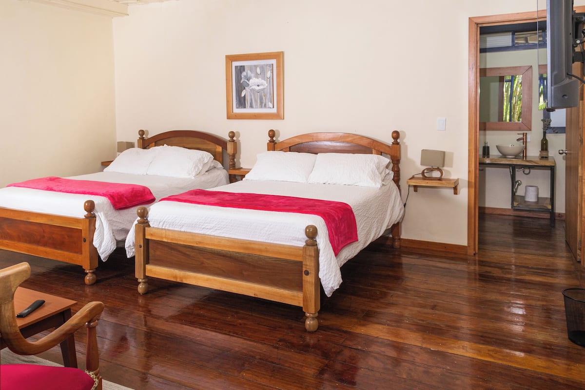 A hotel room with two wooden beds, a wooden chair, a small bedside table with a lamp, and a bathroom visible through an open door.