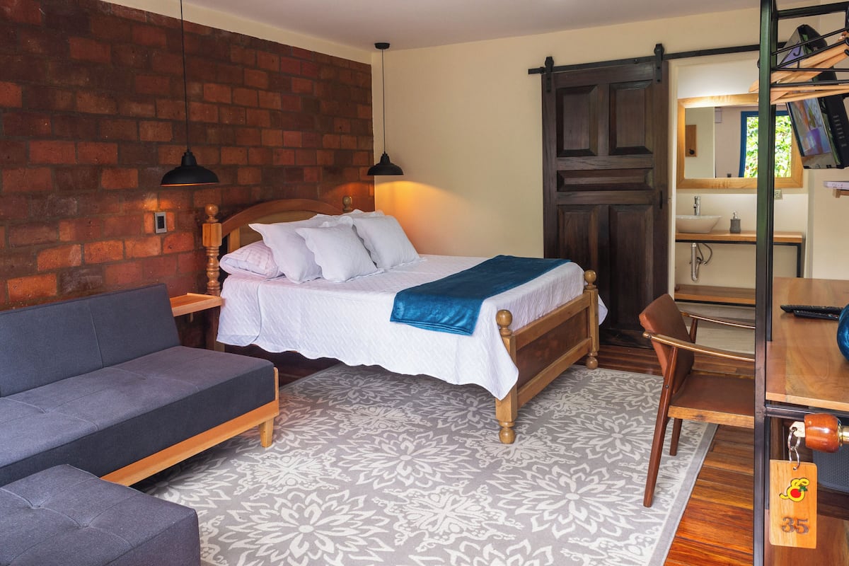 A room with a bed, a grey sofa, a wooden desk, and a brick wall.