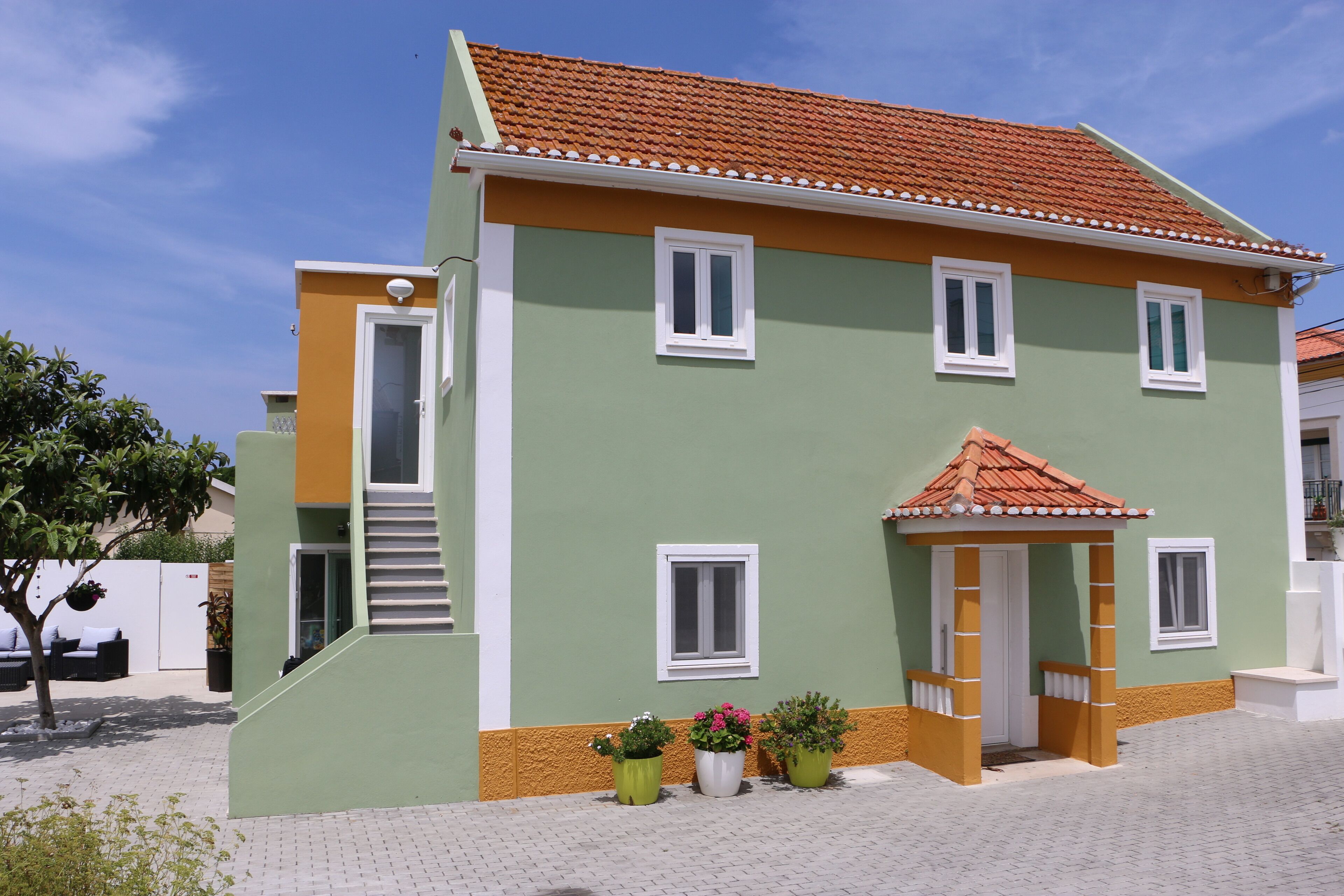 A two-story building with a green and orange facade, a tiled roof, and a small porch with potted plants.