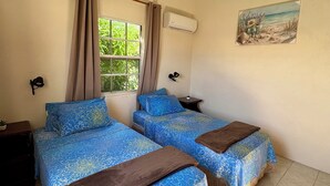 A room with two beds, a window with curtains, an air conditioning unit, a ceiling fan, and a painting on the wall. - Comfy 2- bedroom apartment on Barbados' South Coast, near beach and attractions! (Oistins)