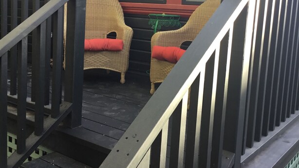 A wooden deck with two wicker chairs and a small table, a staircase with a railing, and a window with blinds.