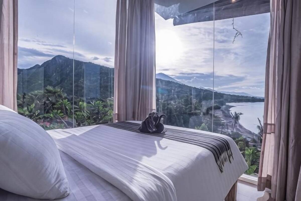 A bed with a view of mountains and a body of water through large windows.