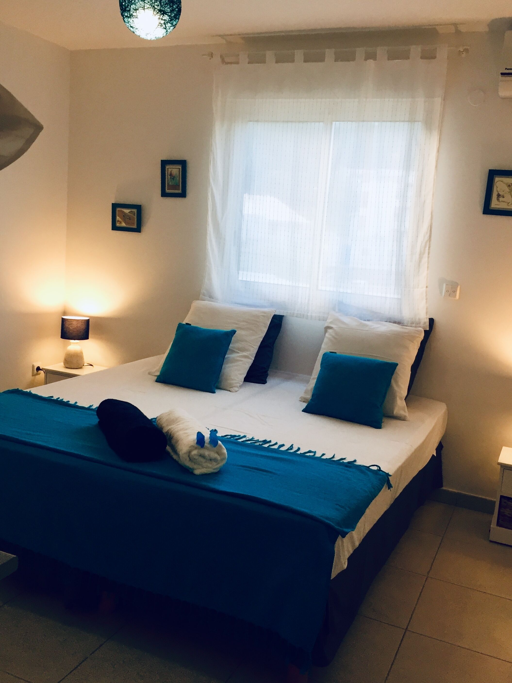 Une chambre à coucher avec un lit, deux lampes de chevet, une fenêtre avec des rideaux légers et un luminaire fixé au mur.
