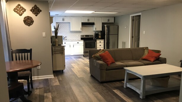 A modern kitchen with white cabinets, a stainless steel refrigerator, and a built-in oven. There is a dining area with a wooden table and chairs, and a living room with a sofa and a coffee table. - Tranquil Escape~Modern 1BD Suite on Greenway (Charlotte)
