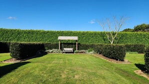 Un jardin bien entretenu, agrémenté d’une pergola en bois et d’une balançoire, entouré de haies soigneusement taillées et surplombé d’un ciel d’un bleu limpide.