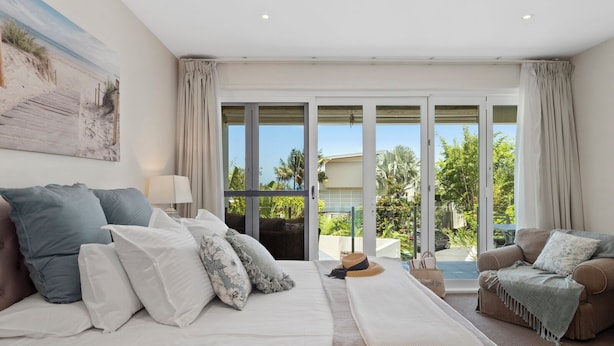 A bedroom with a large bed, a sofa, and a balcony with a view of greenery and a building.