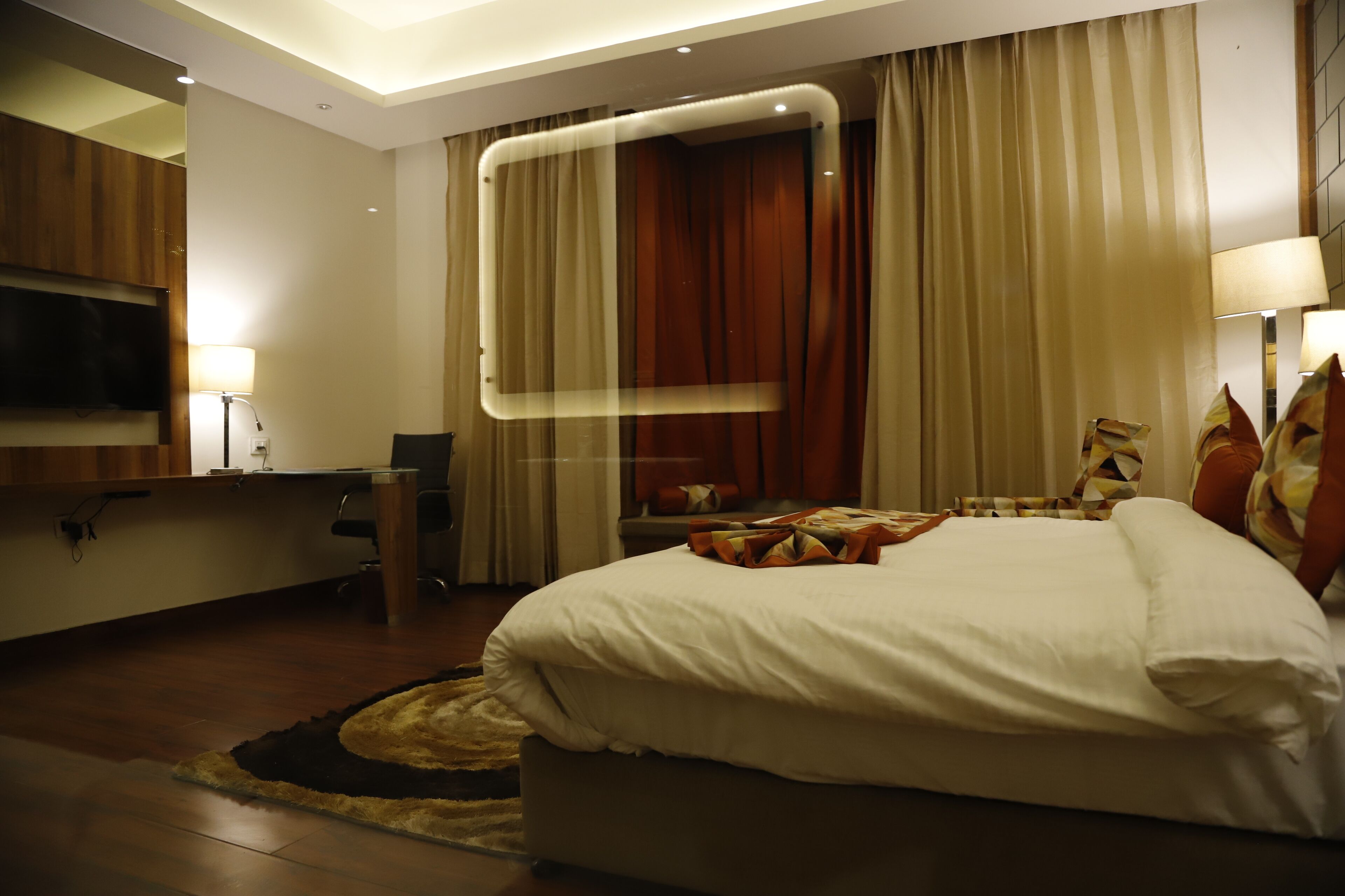 A modern hotel room with a large bed, a desk, a flat-screen TV, and a large window with curtains.
