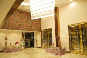 A spacious lobby with a high ceiling, decorative floral wallpaper, and a polished marble floor. There are two reception desks with floral arrangements and red wine glasses.