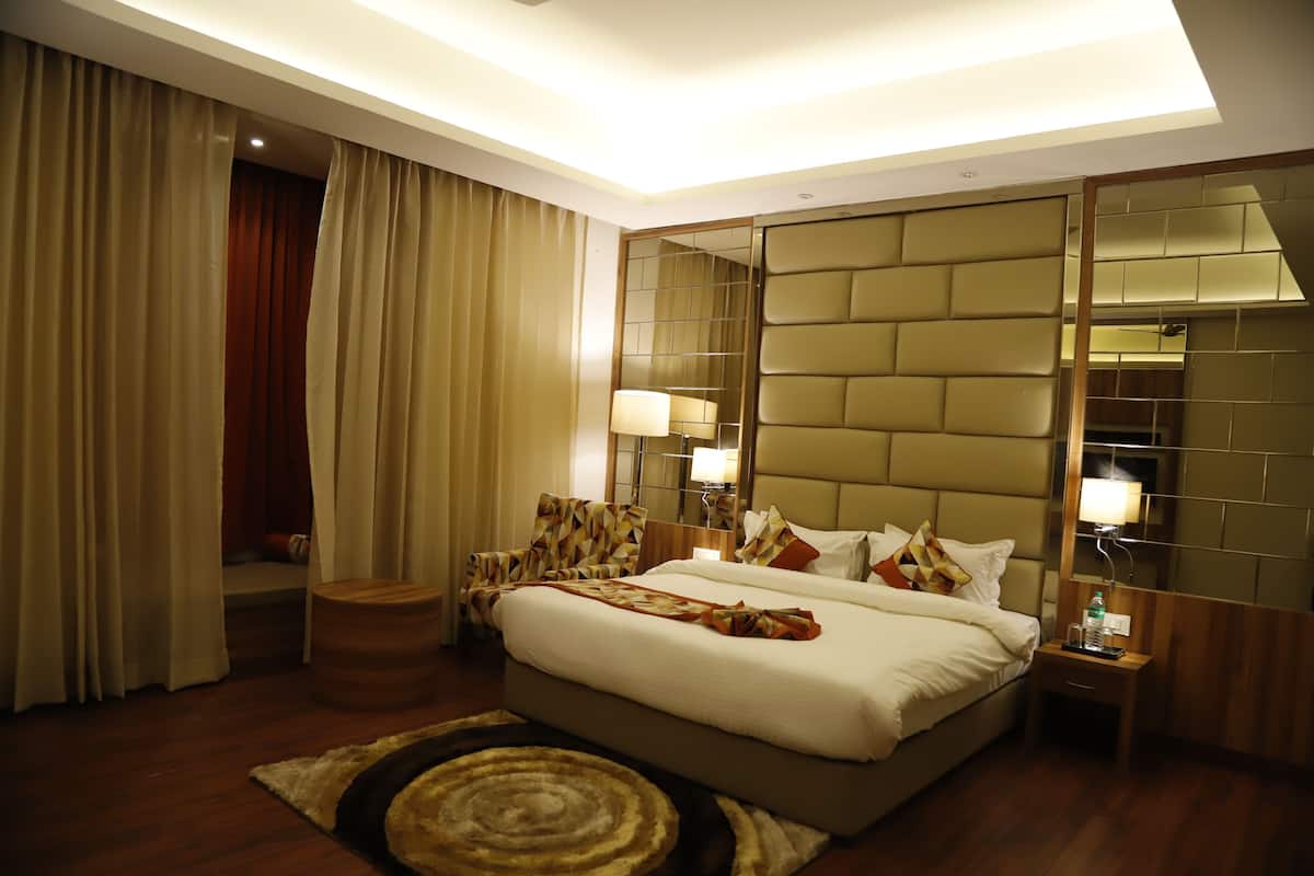 A hotel room with a large bed, bedside lamps, a wooden nightstand, a round wooden stool, and a large mirror on the wall.