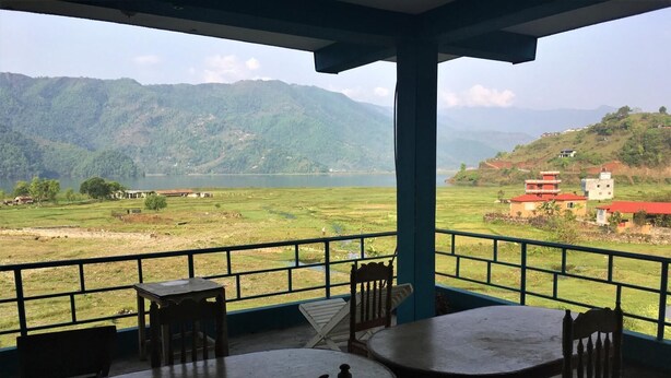 A balcony with a view of a green valley and mountains. There are wooden tables and chairs. - Tranquil Water Guest House Pokhara (Pokhara)