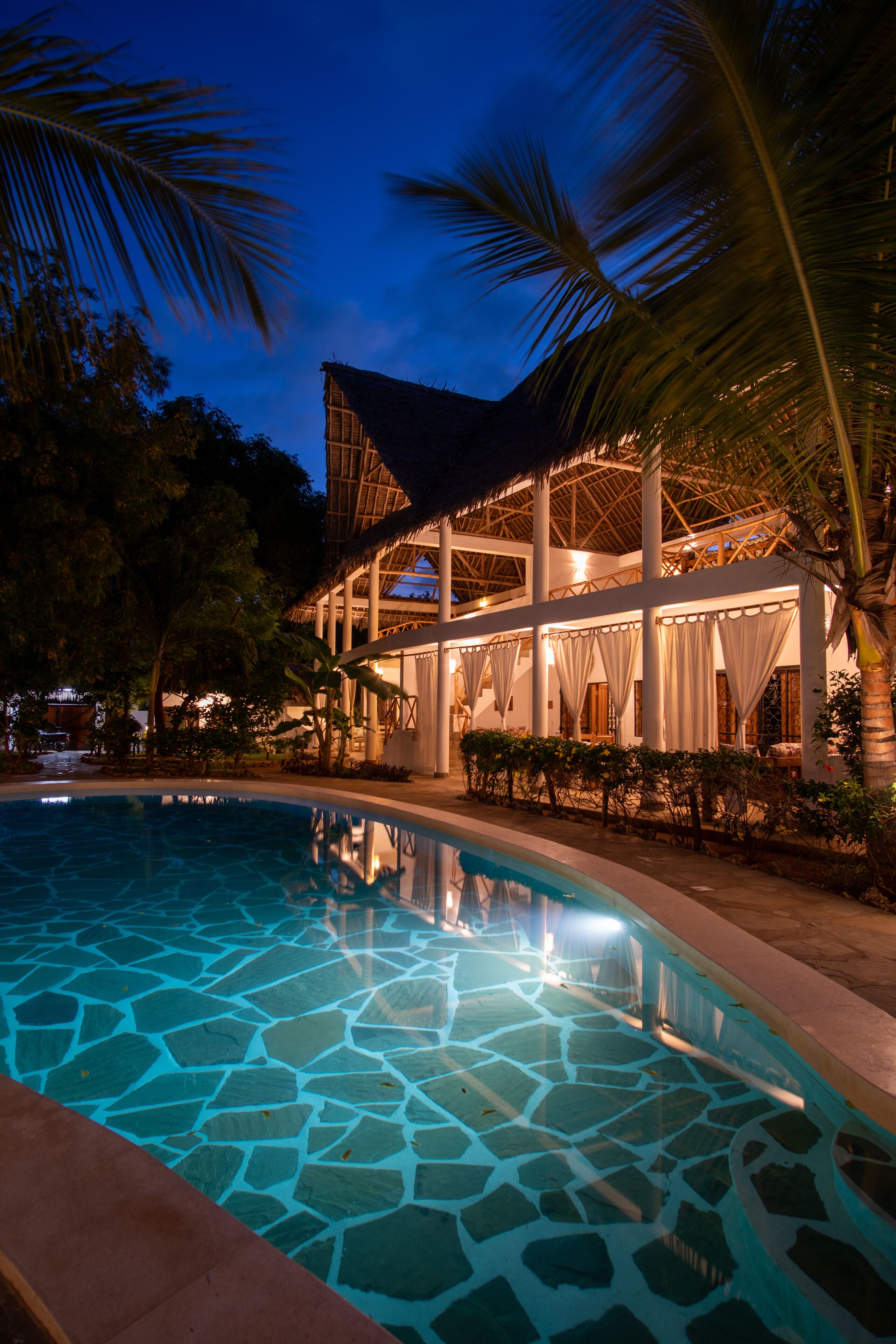 A poolside area with a clear pool, palm trees, and a building with a thatched roof.