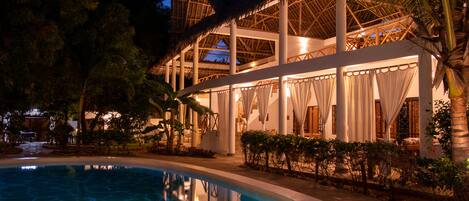 A poolside area with a clear pool, palm trees, and a building with a thatched roof.
