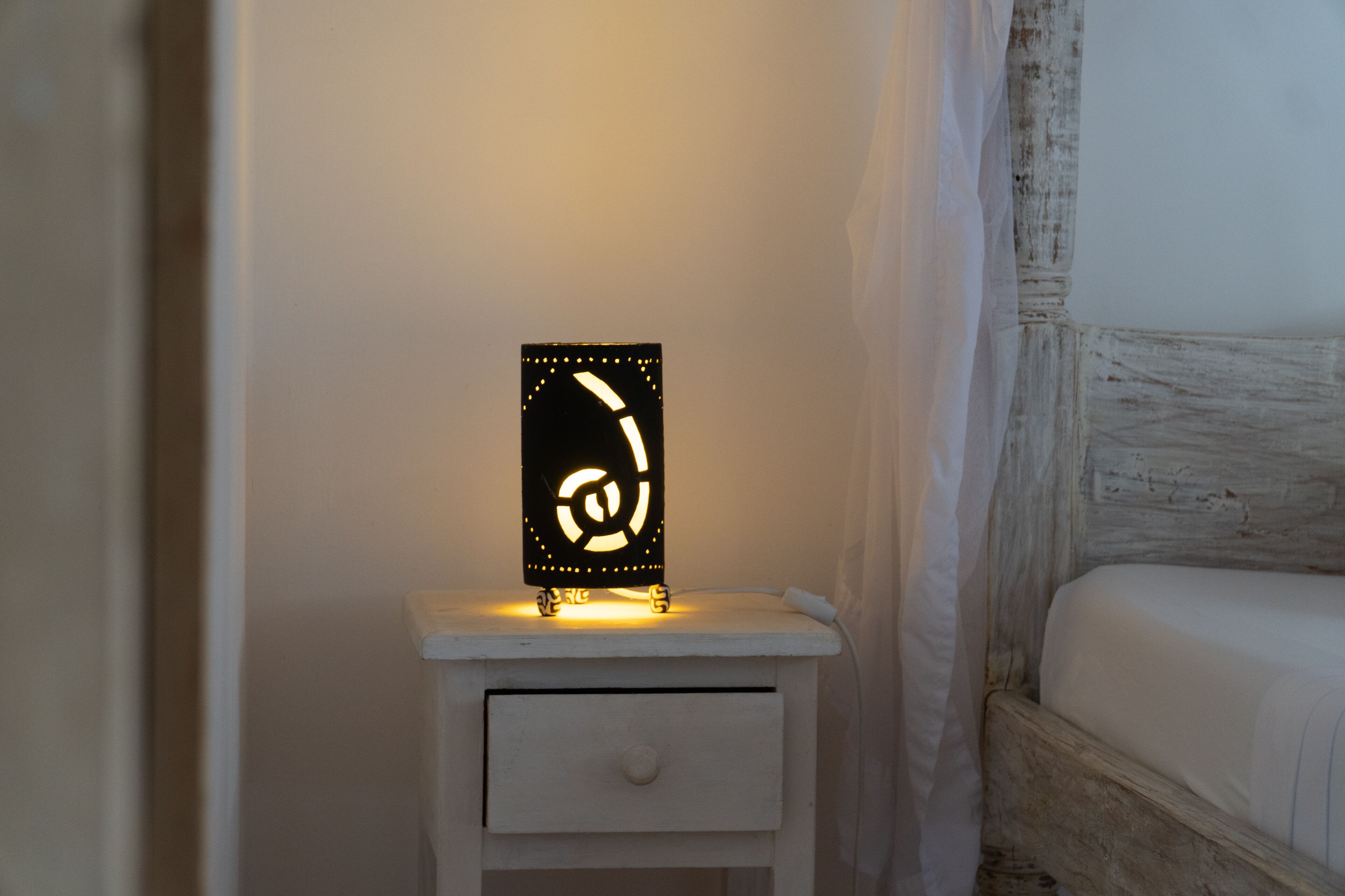 A bedside table with a nightstand lamp, a white drawer, and a bed with a wooden headboard.