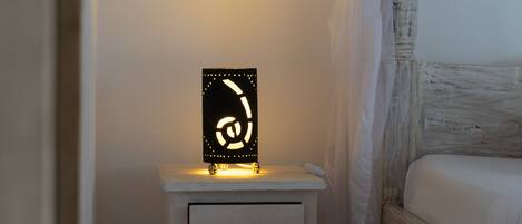 A bedside table with a nightstand lamp, a white drawer, and a bed with a wooden headboard.