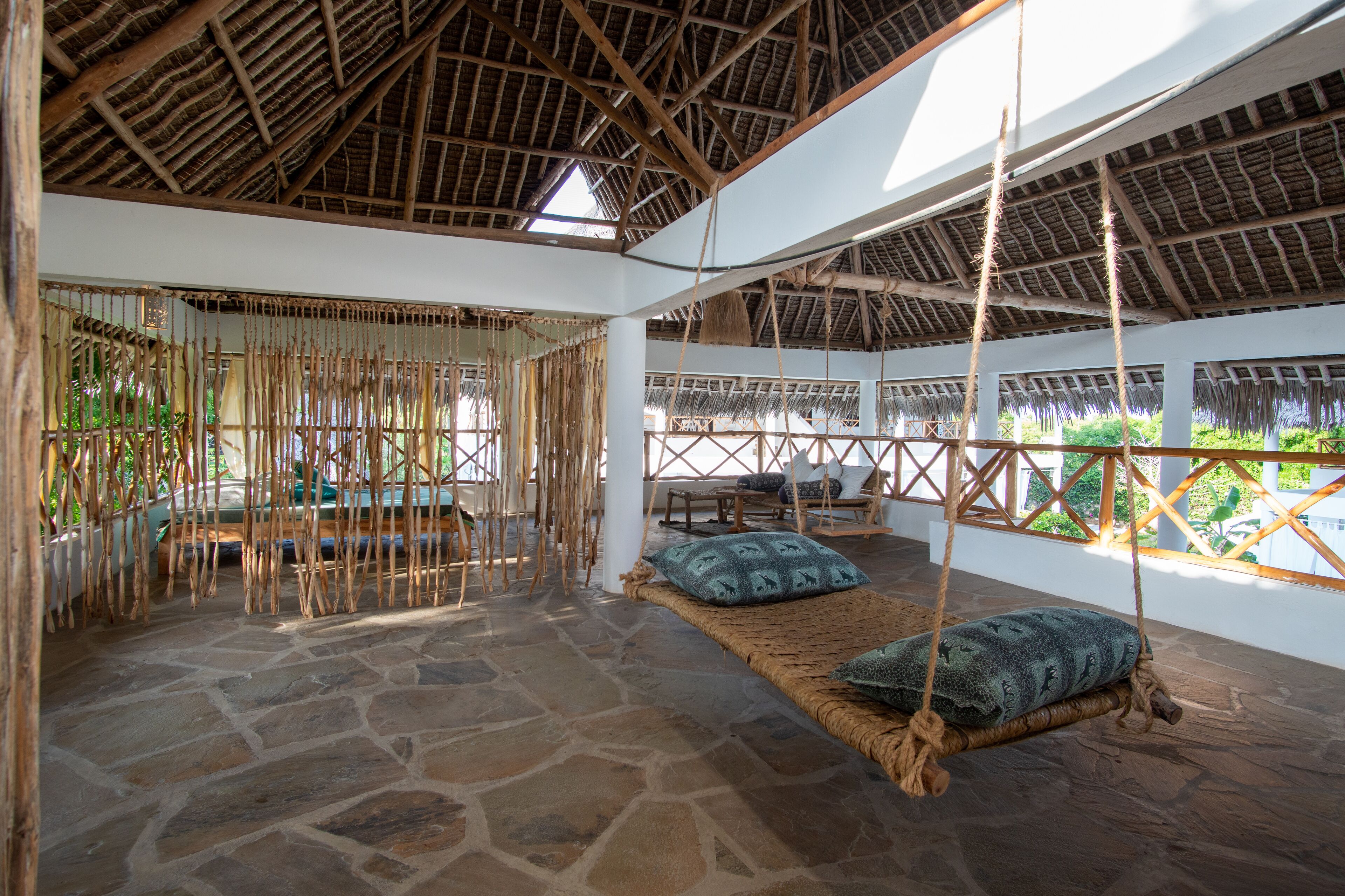 A room with a stone floor, thatched roof, and a hanging swing with cushions.