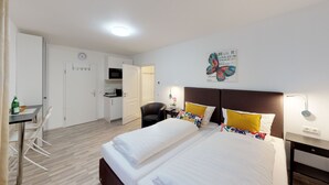 Quarto de hotel com cama, mesas de cabeceira, escrivaninha com cadeira, pequeno espaço de cozinha e relógio na parede.