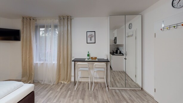 Quarto pequeno e moderno com uma cama, uma secretária com duas cadeiras, uma kitchenette e uma janela grande com cortinas translúcidas.