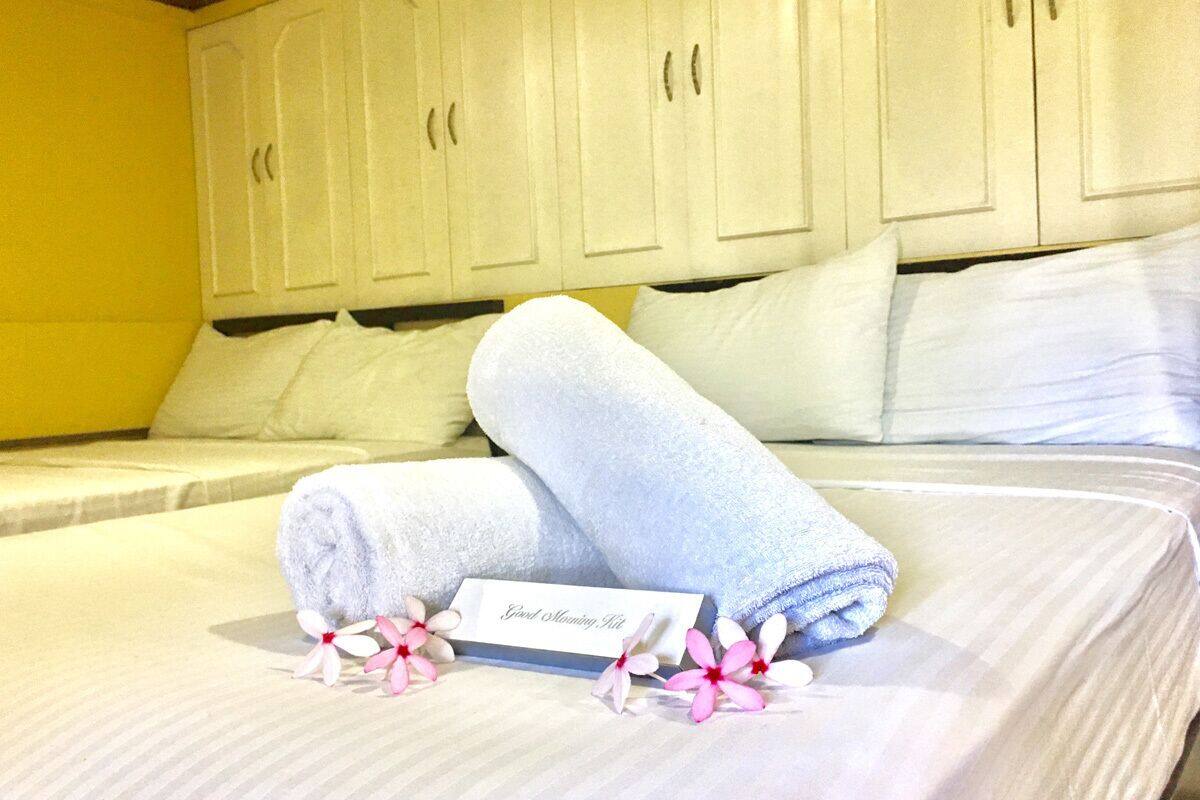 Una cama bien tendida con toallas enrolladas y una tarjeta, almohadas blancas y una pared amarilla.